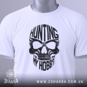 Футболка мисливець дух, біла, складена на поверхні, фон світло-сірий, принт чорного кольору з черепом та написом 'HUNTING MY HOBBY', з етикеткою зеленого кольору.