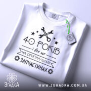 Футболка на оригінальних запчастинах, білого кольору, зображена зверху, на столі, фон світло-сірий, принт чорного кольору з текстом "40 РОКІВ і все це на ОРИГІНАЛЬНИХ ЗАПЧАСТИНАХ" та зображенням ключів і зірочок, з етикеткою зеленого кольору.