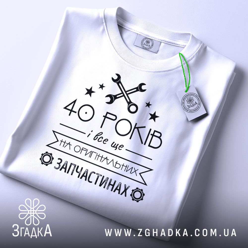 Футболка на оригінальних запчастинах, білого кольору, зображена зверху, на столі, фон світло-сірий, принт чорного кольору з текстом "40 РОКІВ і все це на ОРИГІНАЛЬНИХ ЗАПЧАСТИНАХ" та зображенням ключів і зірочок, з етикеткою зеленого кольору.