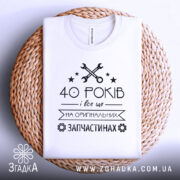 Футболка на оригінальних запчастинах, білого кольору, зображена зверху на плетеній основі, фон світло-сірий, принт чорного кольору з текстом "40 РОКІВ і все це на ОРИГІНАЛЬНИХ ЗАПЧАСТИНАХ" та зображенням ключів і зірочок.