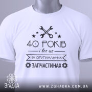 Футболка на оригінальних запчастинах, білого кольору, зображена зверху, на столі, фон світло-сірий, принт чорного кольору з текстом "40 РОКІВ і все це на ОРИГІНАЛЬНИХ ЗАПЧАСТИНАХ" та зображенням ключів і зірочок, з етикеткою зеленого кольору.