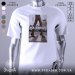 Футболка на завод, біла, зображена з фронтального ракурсу, фон нейтральний, принт з зображенням жіночих ніг у чорних колготках та туфлях на високих підборах, текст "НА ЗАВОД" великими літерами.