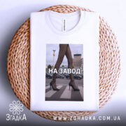 Футболка на завод, біла, зображена зверху на плетеній основі, фон світлий, принт з зображенням жіночих ніг у чорних колготках та туфлях на високих підборах, текст "НА ЗАВОД" великими літерами, видно етикетку на комірці.