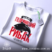 Футболка найголовніший рибак, біла, зображена зверху, на плоскій поверхні, фон світлий, принт червоного кольору з чорним силуетом риби та текстом, на футболці є етикетка.