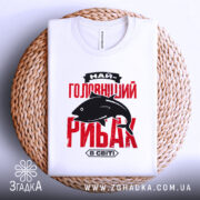 Футболка найголовніший рибак, біла, зображена зверху на плоскій поверхні, фон світлий, футболка лежить на плетеній основі, принт червоного кольору з чорним силуетом риби та текстом.