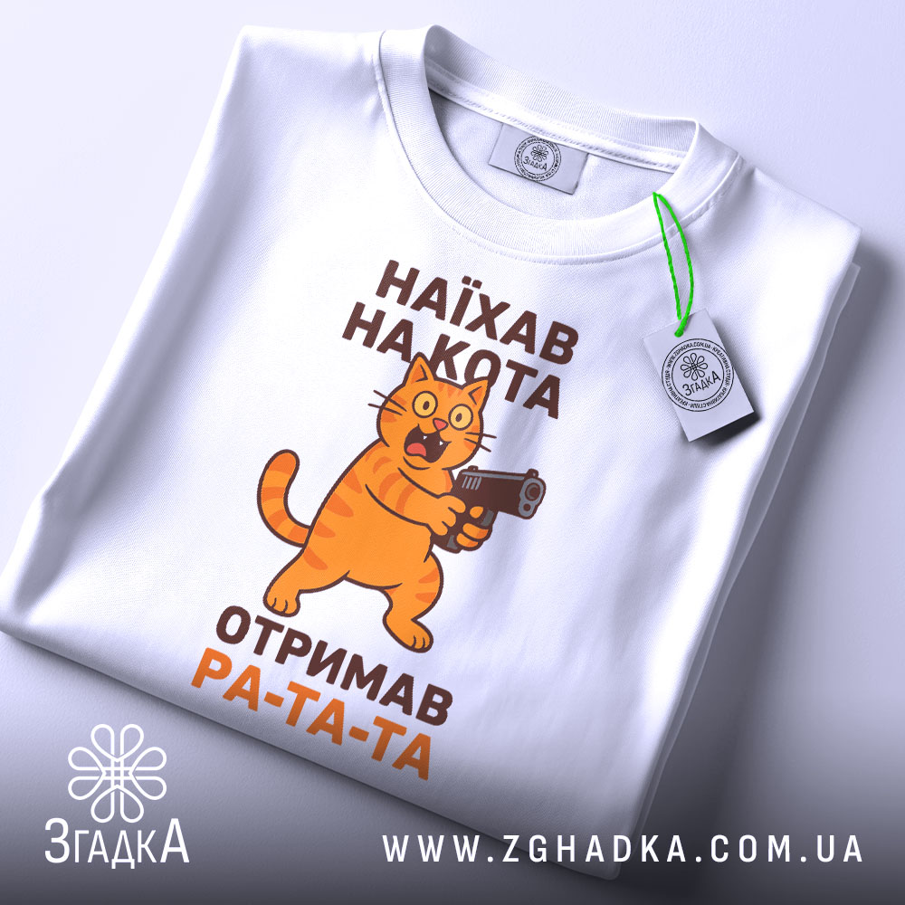 Футболка наїхав на кота, біла, з верхнього ракурсу, на фоні світло-сірого градієнту, принт з жовто-оранжевим котом, що тримає пістолет, текст чорного кольору: 'НАЙХАВ НА КОТА ОТРИМАВ РА-ТА-ТА', з етикеткою зеленого кольору.