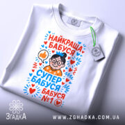 Футболка найкраща бабуся у світі, біла, складена на поверхні, фон світло-сірий, принт з малюнком бабусі в окулярах, з сердечками та написами у яскравих кольорах, на комірці є етикетка.