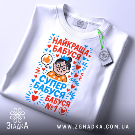 Футболка найкраща бабуся у світі, біла, складена на поверхні, фон світло-сірий, принт з малюнком бабусі в окулярах, з сердечками та написами у яскравих кольорах, на комірці є етикетка.