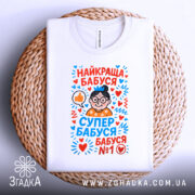 Футболка найкраща бабуся у світі, біла, складена на плетеній основі, фон білий, принт з малюнком бабусі в окулярах, з сердечками та написами у яскравих кольорах.