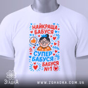 Футболка найкраща бабуся у світі, біла, складена на поверхні, фон світло-сірий, принт з малюнком бабусі в окулярах, з сердечками та написами у яскравих кольорах, на комірці є етикетка.