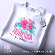 Футболка найкраща донечка білого кольору, зображена в горизонтальному положенні, фон світлий, на комірці є етикетка, принт з рожевими квітами та написом "найкраща донечка у світі" в яскравих кольорах.