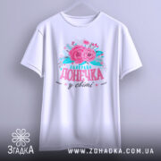 Футболка найкраща донечка білого кольору, підвішена на вішалці, фон світло-сірий, принт з рожевими квітами та написом "найкраща донечка у світі" в яскравих кольорах.