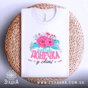 Футболка найкраща донечка білого кольору, зображена в горизонтальному положенні на плетеній основі, фон світлий, принт з рожевими квітами та написом "найкраща донечка у світі" в яскравих кольорах.