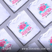 Футболка найкраща донечка білого кольору, зображена на манекені, фон світло-сірий, принт з рожевими квітами та написом "найкраща донечка у світі" в яскравих кольорах.