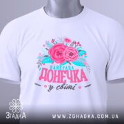 Футболка найкраща донечка білого кольору, зображена в горизонтальному положенні, фон світлий, на комірці є етикетка, принт з рожевими квітами та написом "найкраща донечка у світі" в яскравих кольорах.