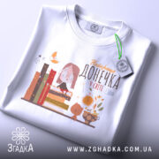 Футболка найкраща донечка, білого кольору, зображена зверху на плоскій поверхні, фон світло-сірий, принт містить малюнок дівчинки в окулярах, що сидить на книжках, поряд з котом і квітами, з етикеткою на шнурку.