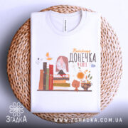 Футболка найкраща донечка, білого кольору, зображена зверху на плоскій поверхні, фон світло-сірий, принт містить малюнок дівчинки в окулярах, що сидить на книжках, поряд з котом і квітами, на плетеній основі.