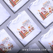 Футболка найкраща донечка, білого кольору, зображена з фронтального ракурсу на манекені, фон світло-сірий, принт містить малюнок дівчинки в окулярах, що сидить на книжках, поряд з котом і квітами.