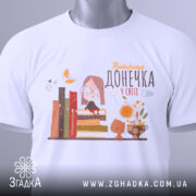 Футболка найкраща донечка, білого кольору, зображена зверху на плоскій поверхні, фон світло-сірий, принт містить малюнок дівчинки в окулярах, що сидить на книжках, поряд з котом і квітами, з етикеткою на шнурку.