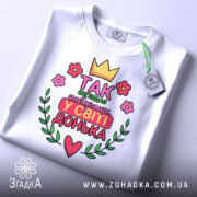 Футболка найкраща донька, біла, складена на поверхні, фон світло-сірий, принт з короною, квітами та написом 'Так виглядає найкраща у світі донька', з зеленим ярликом.