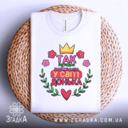 Футболка найкраща донька, біла, складена на плетеній кошику, фон світло-сірий, принт з короною, квітами та написом 'Так виглядає найкраща у світі донька'.