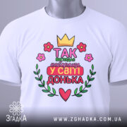 Футболка найкраща донька, біла, складена на поверхні, фон світло-сірий, принт з короною, квітами та написом 'Так виглядає найкраща у світі донька', з зеленим ярликом.