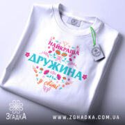 Футболка найкраща дружина білого кольору, складена на поверхні, фон світло-сірий, принт яскравий з написом 'НАЙКРАЩА ДРУЖИНА у світі', на футболці є етикетка з зеленим шнурком.