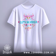 Футболка найкраща дружина білого кольору, зображена на вішалці, фон світло-сірий, принт яскравий з написом 'НАЙКРАЩА ДРУЖИНА у світі', футболка має вільний крій.