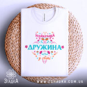 Футболка найкраща дружина білого кольору, складена на плетеній кошику, фон світло-сірий, принт яскравий з написом 'НАЙКРАЩА ДРУЖИНА у світі', видна етикетка на горловині.