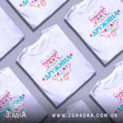 Футболка найкраща дружина білого кольору, зображена на манекені з правого боку, фон світло-сірий, принт містить яскраві кольорові елементи з написом 'НАЙКРАЩА ДРУЖИНА у світі'.