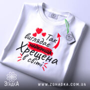 Футболка найкраща хрещена, білого кольору, зображена зверху, на фоні світло-сірого кольору, принт з червоними сердечками та текстом "Так виглядає найкраща Хрещена в світі", на футболці також є етикетка.