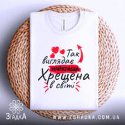 Футболка найкраща хрещена, білого кольору, зображена зверху на плетеній основі, фон білий, принт містить червоні сердечка та текст "Так виглядає найкраща Хрещена в світі".