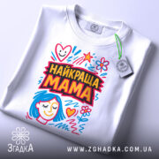 Футболка найкраща мама, біла, складена на столі, фон світло-сірий, принт з яскравими кольорами, з малюнком обличчя жінки з блакитним волоссям та сердечками, з етикеткою на шнурку.