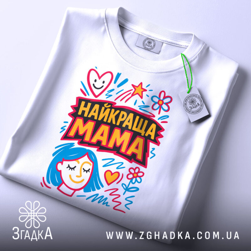 Футболка найкраща мама, біла, складена на столі, фон світло-сірий, принт з яскравими кольорами, з малюнком обличчя жінки з блакитним волоссям та сердечками, з етикеткою на шнурку.