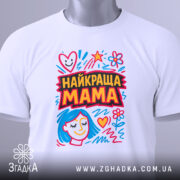 Футболка найкраща мама, біла, складена на столі, фон світло-сірий, принт з яскравими кольорами, з малюнком обличчя жінки з блакитним волоссям та сердечками, з етикеткою на шнурку.