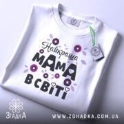 Футболка найкраща мама, білого кольору, зображена зверху на плоскій поверхні, фон світло-сірий, принт з квітами та написом "Найкраща мама в світі" в чорному та фіолетовому кольорах, з етикеткою на комірці.