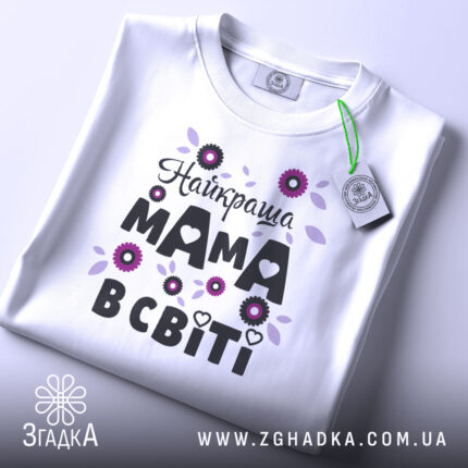 Футболка найкраща мама, білого кольору, зображена зверху на плоскій поверхні, фон світло-сірий, принт з квітами та написом "Найкраща мама в світі" в чорному та фіолетовому кольорах, з етикеткою на комірці.