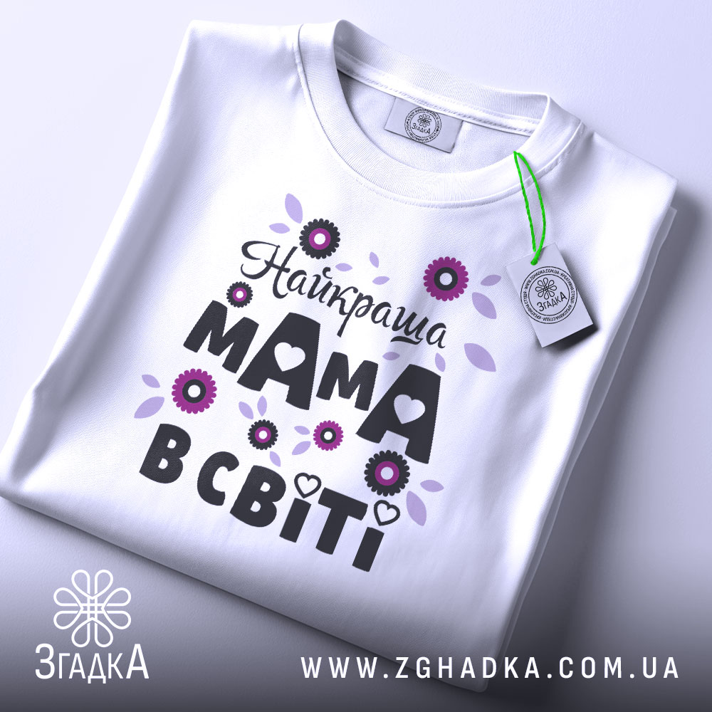 Футболка найкраща мама, білого кольору, зображена зверху на плоскій поверхні, фон світло-сірий, принт з квітами та написом "Найкраща мама в світі" в чорному та фіолетовому кольорах, з етикеткою на комірці.