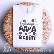 Футболка найкраща мама, білого кольору, зображена зверху на плоскій поверхні, фон світло-сірий, принт з квітами та написом "Найкраща мама в світі" в чорному та фіолетовому кольорах, розташована на плетеній основі.