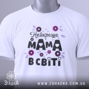 Футболка найкраща мама, білого кольору, зображена зверху на плоскій поверхні, фон світло-сірий, принт з квітами та написом "Найкраща мама в світі" в чорному та фіолетовому кольорах, з етикеткою на комірці.