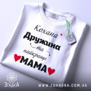 Футболка найкраща мама, біла, складена на поверхні, фон світло-сірий, принт чорний з червоними сердечками, текст українською мовою, на ярлику зелена стрічка.