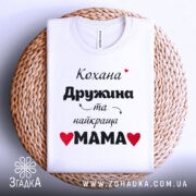 Футболка найкраща мама, біла, складена на кошику, фон світло-сірий, принт чорний з червоними сердечками, текст українською мовою.