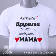 Футболка найкраща мама, біла, складена на поверхні, фон світло-сірий, принт чорний з червоними сердечками, текст українською мовою, на ярлику зелена стрічка.