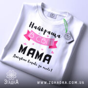 Футболка найкраща мама, білого кольору, складена на столі, фон світло-сірий, принт з текстом "Найкраща в світі МАМА" з рожевими елементами та сердечками, на комірці є етикетка.