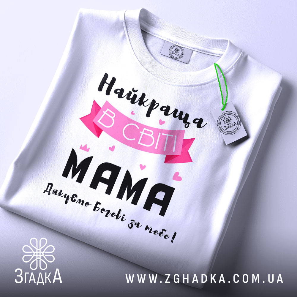 Футболка найкраща мама, білого кольору, складена на столі, фон світло-сірий, принт з текстом "Найкраща в світі МАМА" з рожевими елементами та сердечками, на комірці є етикетка.