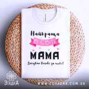 Футболка найкраща мама, білого кольору, складена на плетеній основі, фон світло-сірий, принт містить текст "Найкраща в світі МАМА" з рожевими елементами та сердечками.