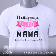 Футболка найкраща мама, білого кольору, складена на столі, фон світло-сірий, принт з текстом "Найкраща в світі МАМА" з рожевими елементами та сердечками, на комірці є етикетка.