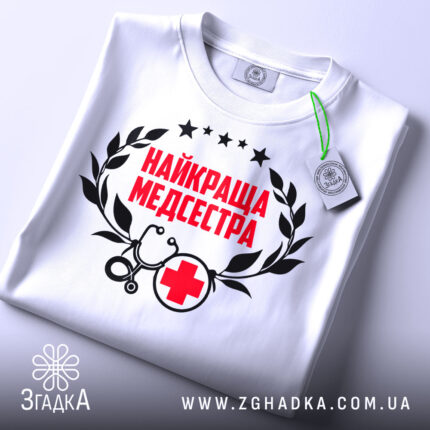 Футболка найкраща медсестра, білого кольору, зображена зверху на плоскій поверхні, фон світло-сірий, принт містить червоний текст "НАЙКРАЩА МЕДСЕСТРА" з чорними елементами, на футболці є етикетка з зеленим шнурком.