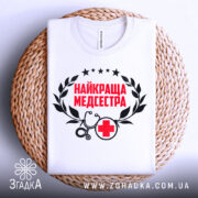 Футболка найкраща медсестра, білого кольору, зображена зверху на плоскій поверхні, фон світло-сірий, принт містить червоний текст "НАЙКРАЩА МЕДСЕСТРА" з чорними елементами, футболка розташована на плетеній основі.