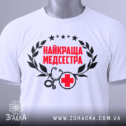 Футболка найкраща медсестра, білого кольору, зображена зверху на плоскій поверхні, фон світло-сірий, принт містить червоний текст "НАЙКРАЩА МЕДСЕСТРА" з чорними елементами, на футболці є етикетка з зеленим шнурком.